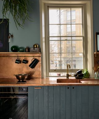 kitchen countertop trends, blue/green kitchen with copper countertop and backsplash, cream painted window frame, brass tap, open shelf, artwork