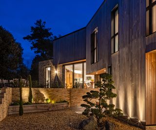 Outside of contemporary home clad in wood with uplights illuminating the outside