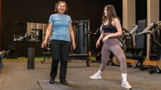 Two women in a gym. A younger woman demonstrates a leg position while the older woman looks forward smiling