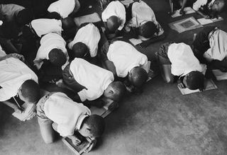 Black and white photograph of several schoolchildren bent over on the floor to write, some write on paper others write on chalkboards.