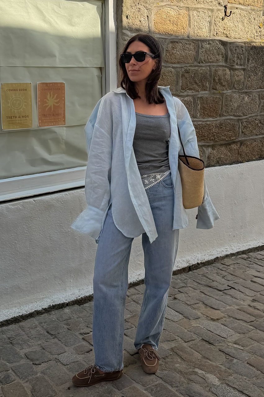 a woman wearing a blue oversized button-down shirt over a gray tnak top, blue jeans, a white lace scarf on her waist, a straw woven bag, and boat shoes