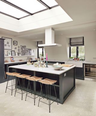 Grey kitchen cabinetry and island with rattan barstools in a white kitchen scheme with pale stone flooring.