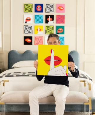 Jonathan Adler sitting on a bench in front of a bed holding a bright yellow print