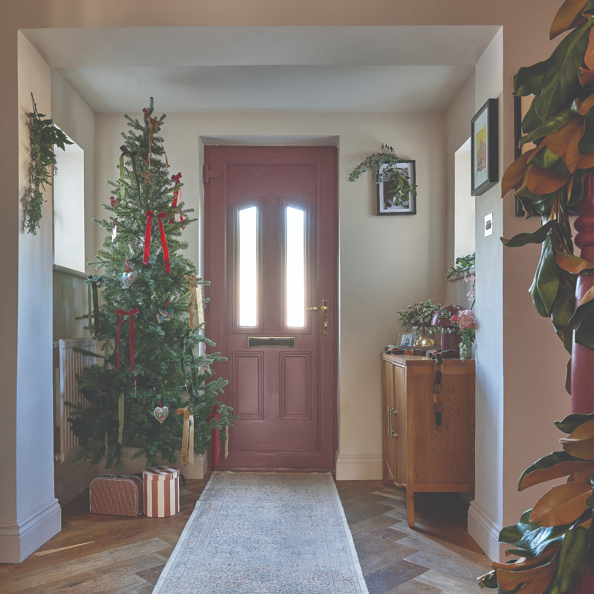 A hallway with a pink front door decorated for Christmas with a slim Christmas tree adorned with bows