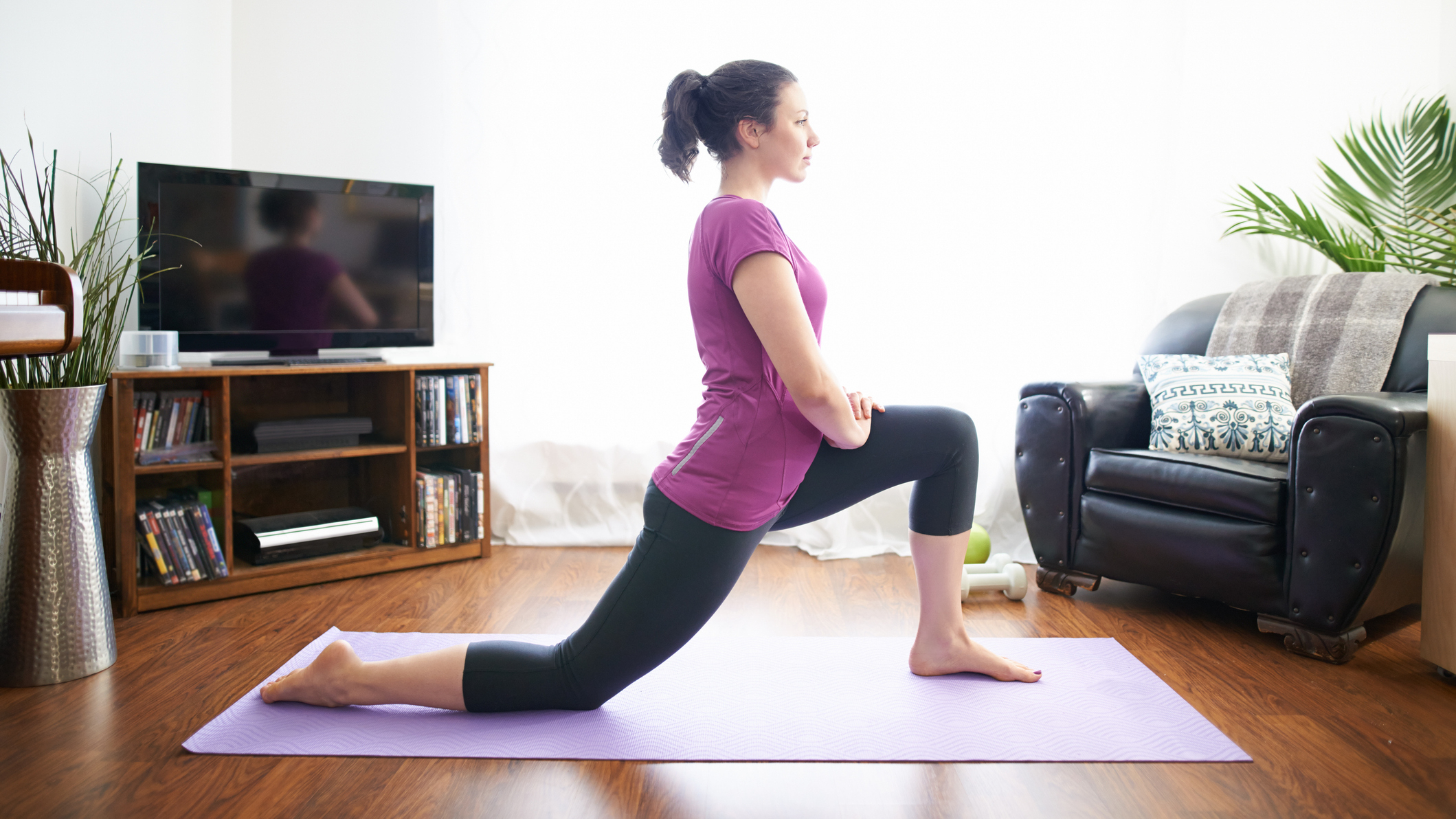 Woman stretching her hip flexors in a low lunge position between a couch and a TV