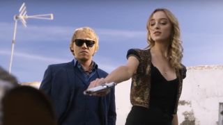 Rupert Grint in sunglasses standing calmly as Phoebe Dynevor holds a pistol on Snatch