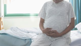 photo shows a pregnant woman in a paper gown sitting on a hospital bed with her hands on her stomach