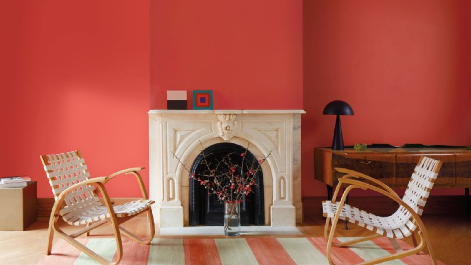Living room with bright red walls, marble fireplace at the centre with a large modern painting propped up on it, two woven lounge chairs, a striped red and white rug, and a midcentury console in the corner with a black lamp on top of it.