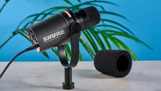 the shure mv7i, a black microphone with a black muff, touch LED pad, XLR connection and TRS input and USB-C input at the rear, photograhped against a blue tom's guide background with a pair of over-ear wired headphones