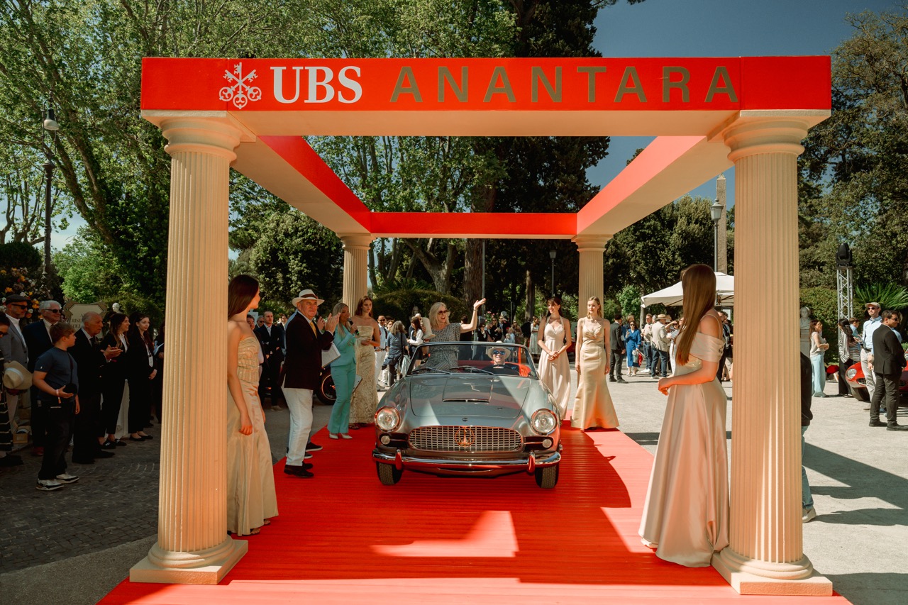 A silver classic Maserati drives beneath a red UBS and Anantara archway at Anantara Concorso Roma, flanked by elegantly dressed hosts as spectators look on.