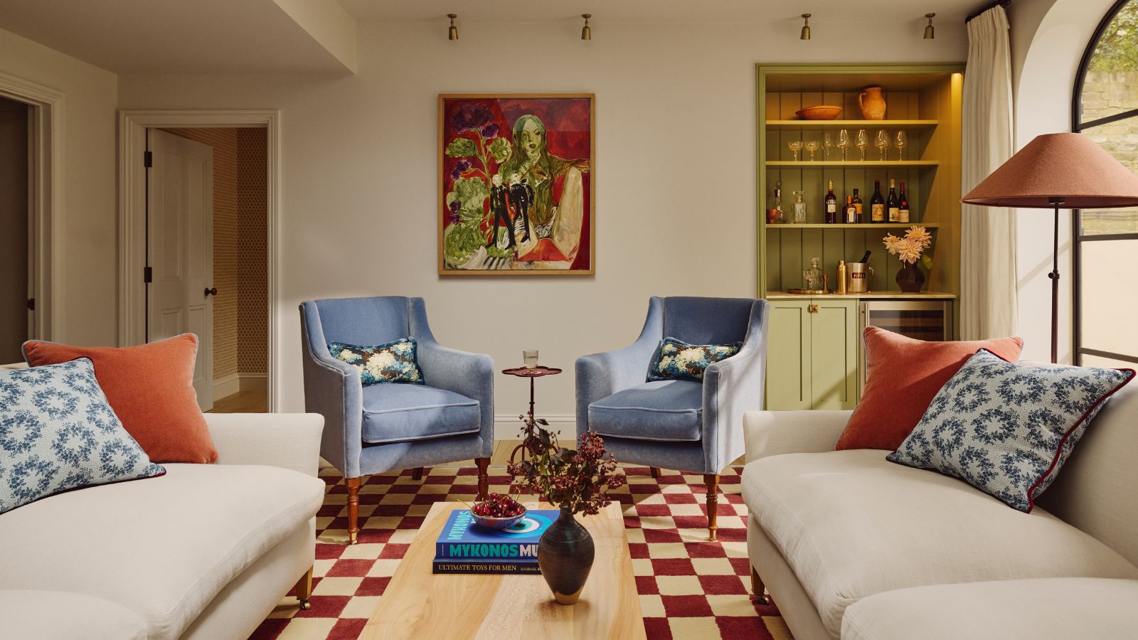neutral living room with bold art, two white sofas, blue armchairs, red checkerboard rug and a green home bar in the corner