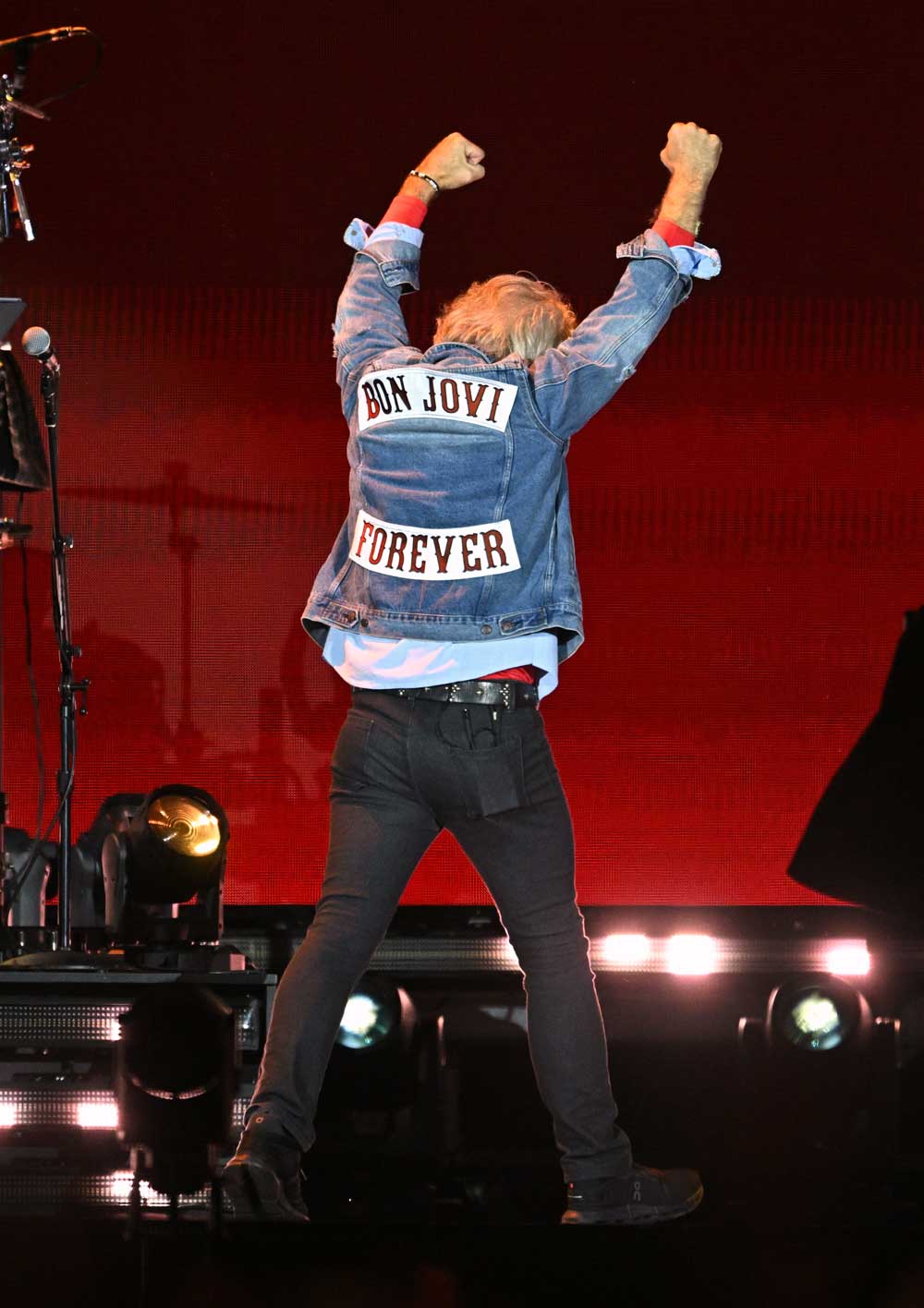 Jon Bon Jovi onstage, arms aloft, wearing a 'Bon Jovi Forever' denim, jacket