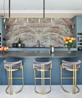 A dusky blue kitchen island with three gray-blue and gold bar stools in front, large gold pendant light above it, and a brown marble splash with blue hues