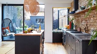 a kitchen with dark kitchen units with an island with wood countertop and exposed brick