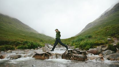 A man crosses a stream in the mountains by walking across big rocks.