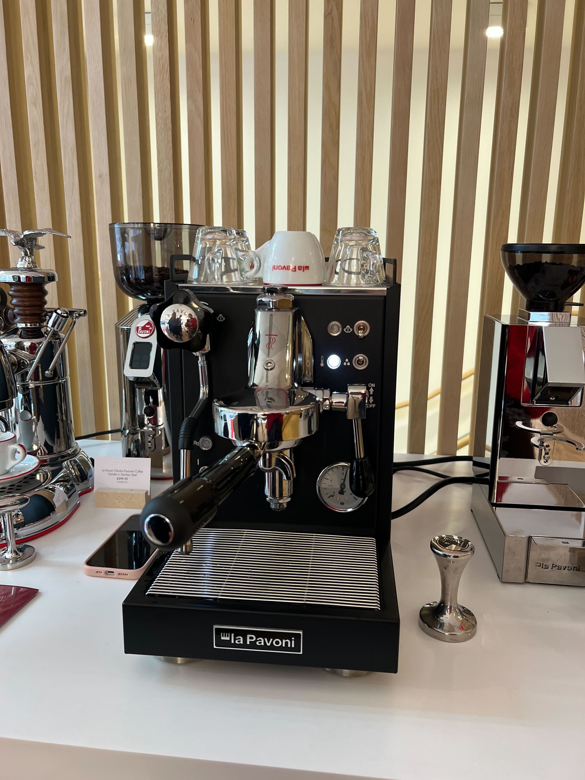 A black La Pavoni Mini Cellini Semi-Professional Domestic Coffee Machine on a white countertop with other machine around it.