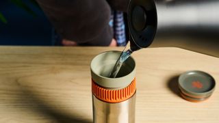 Pouring water into the Wacaco Pipamoka portable coffee maker in front of a blue background.