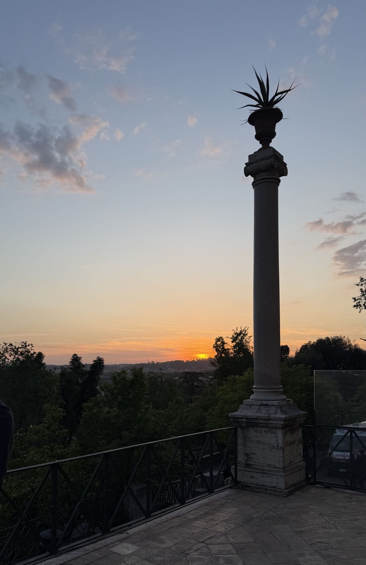 The sun sets beyond the trees at Anantara Concorso Roma, silhouetting a stone column topped with a decorative urn against a pastel evening sky.