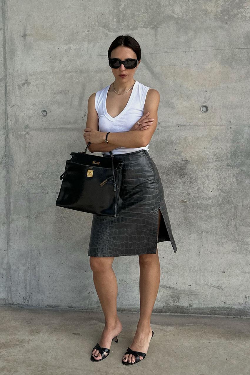 A woman wearing a white V-neck tank top, black croc-embossed skirt, black sandals, and a black Hermes bag.