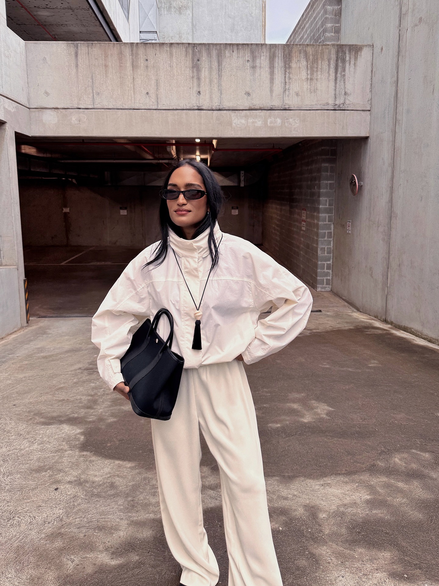 Div Ravindran in a white windbreaker, white pants, black tassel necklace, and black handbag