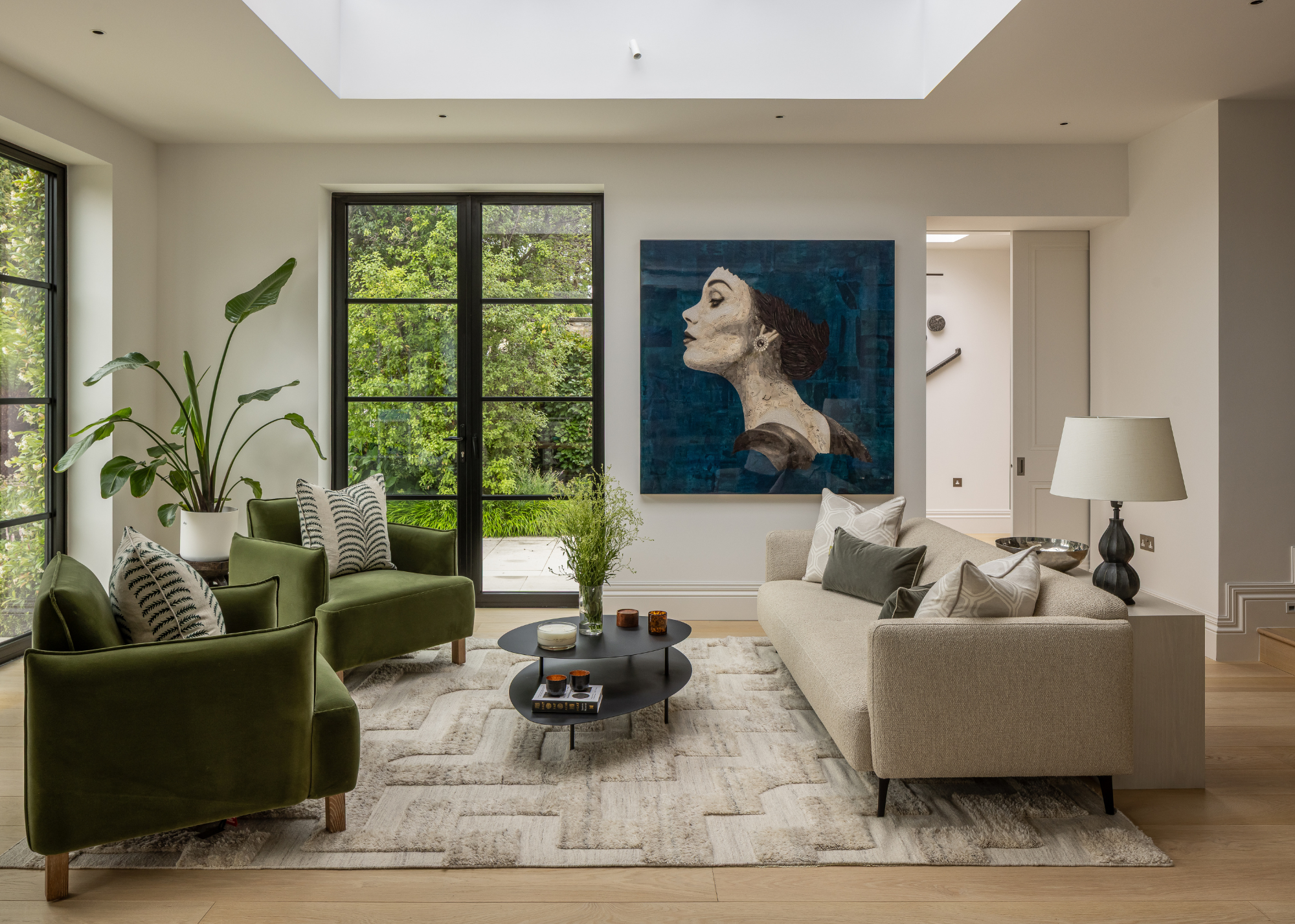 A modern living space with a green and beige sofa facing each other with a black two-layered thin coffee table in the center styled with a vase of foliage, books and candles; and there is a large artwork of the side of a girl's face on the wall next to black-framed doors leading to the garden