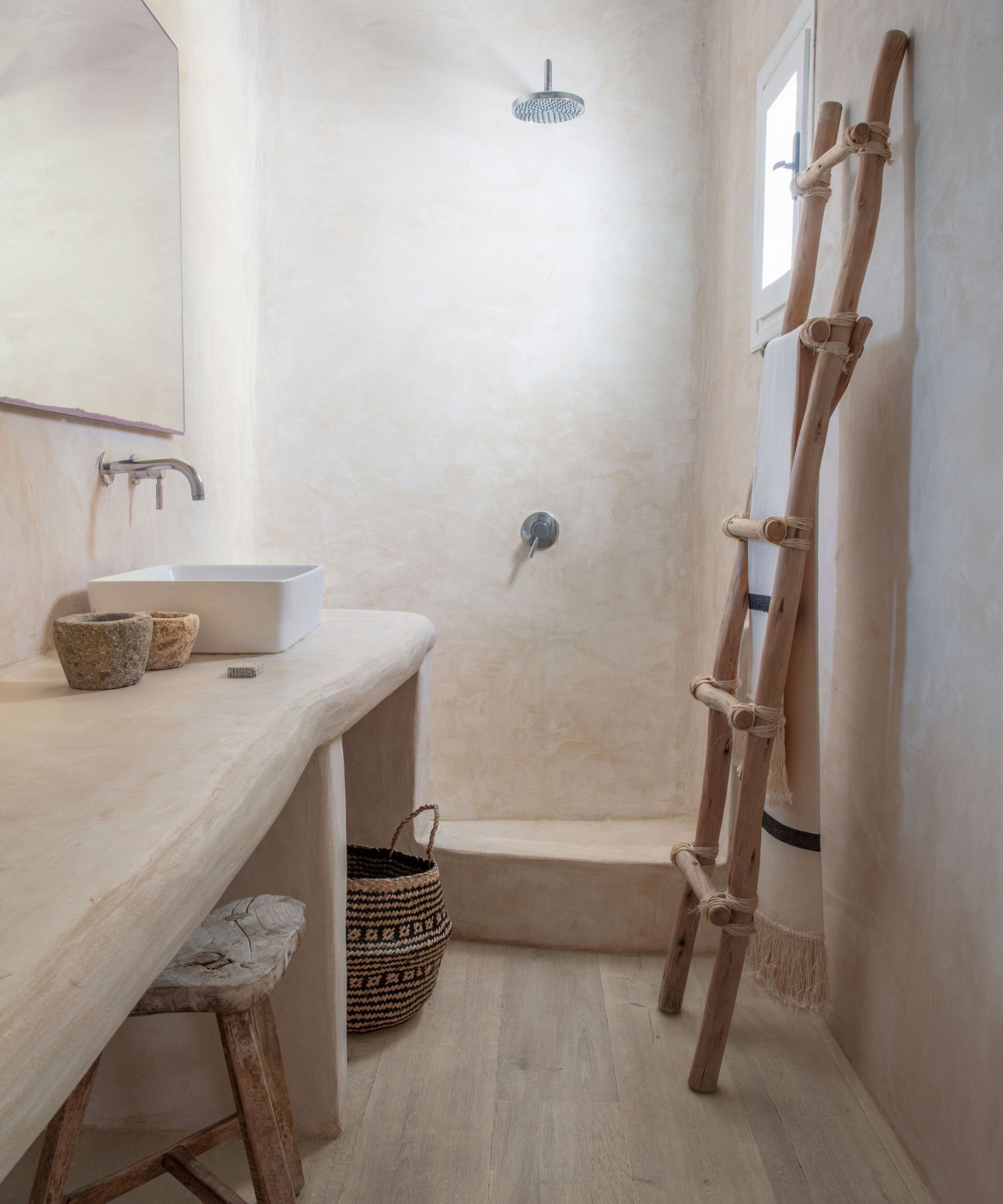 A rustic, minimalist bathroom entirely finished in smooth sandy plaster, walls, floor and vanity included. A white rectangular vessel basin sits on a raw plaster shelf, with a driftwood branch ladder leaning against the wall holding a towel, beside a simple open rain shower.