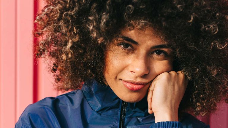 A young woman is smiling directly at the camera, she has curly hair and is leaning on her hand