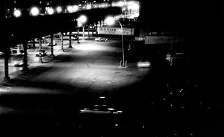 Dark black and white image showing cars with headlights on travelling below a pier-like structure