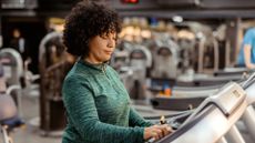 woman in a green long sleeved top on a treadmill and adjusting the settings; the camera shot is from the waist up with a gym setting behind her.