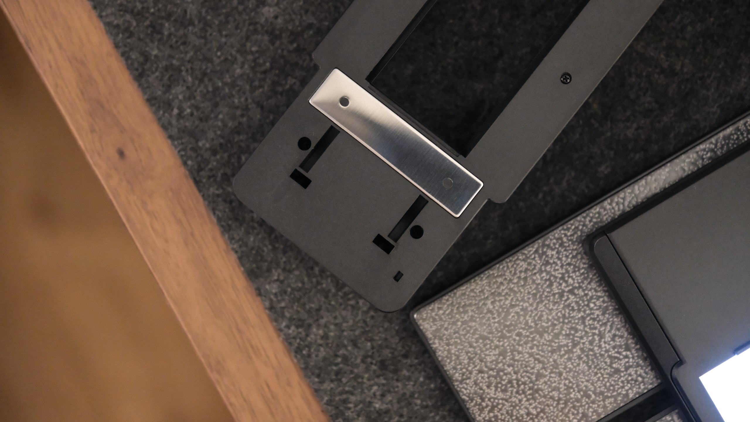A close-up showing the underside of a black plastic film scanning accessory, featuring a rectangular silver metal plate secured with small screws, resting flat on a grey surface.