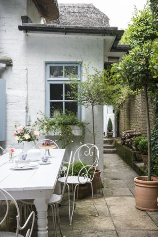 a small cottage garden patio with a white dining table and chairs, with a path leading down to the front door, and windows painted light blue