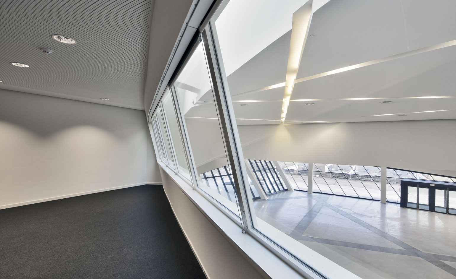 Room with a grey carpet, view from the large internal windows, down from the first floor onto the lower foyet and outside world via the large glass windows and black frame entrance doors at the front of the centre