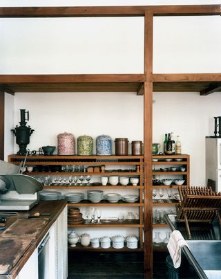A detail of the kitchen. A wooden shelf is set against the far wall and is filled with dishes, glasses, and bottles of oil. The sink is to the right, and cabinets with plenty of counter space to the left.