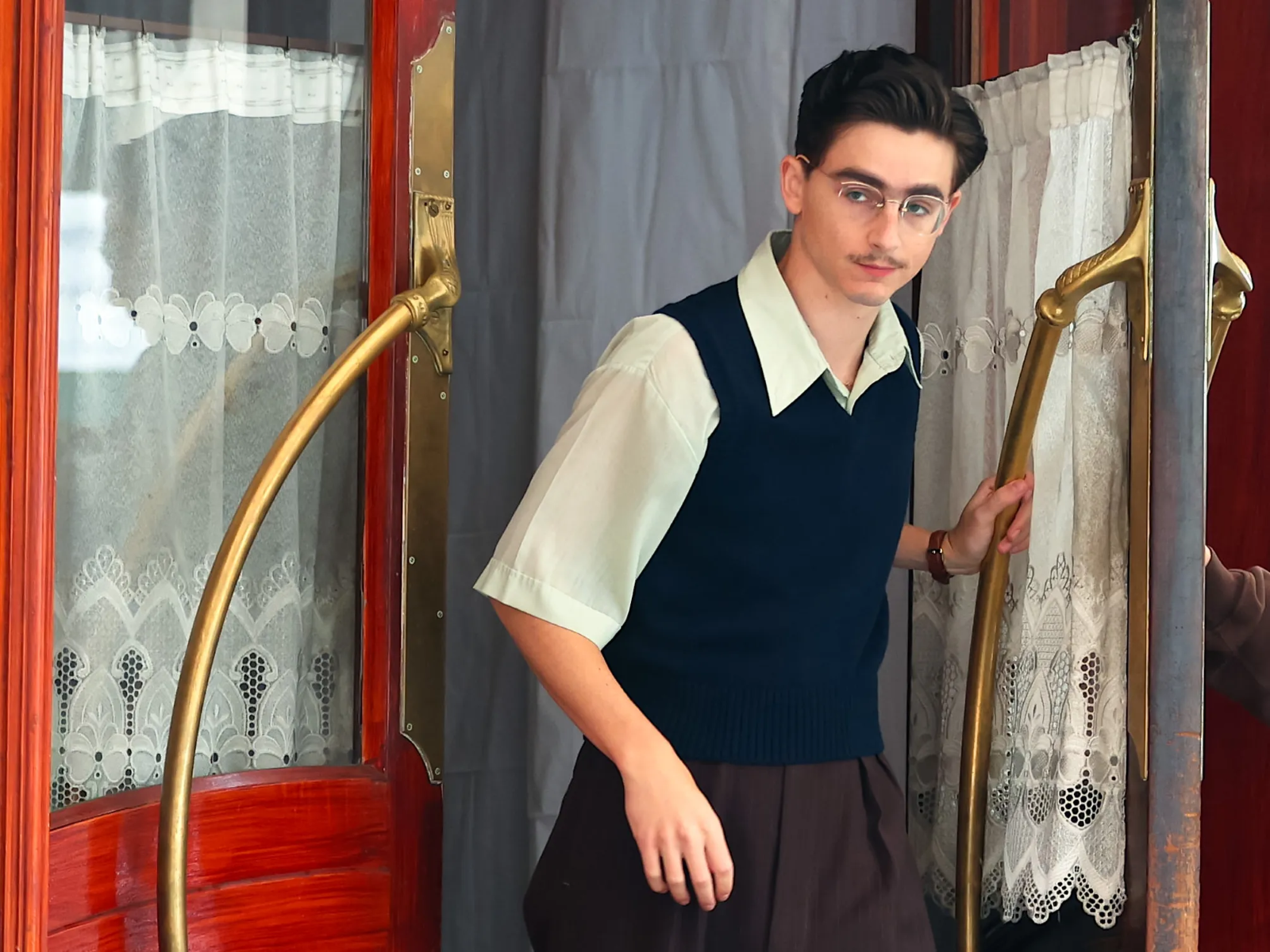 an image of Timothee Chalamet from the upcoming film, Marty Supreme, he is wearing glasses and a sweater vest, exiting from a building with red doors