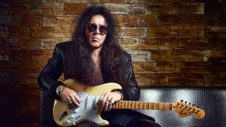 Yngwie Malmsteen holding his guitar sitting on a couch