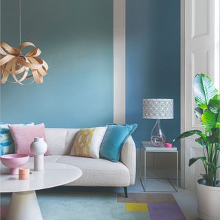 A living room with a colour-blocked blue wall and a sculptural wooden pendant light hanging above the round white coffee table