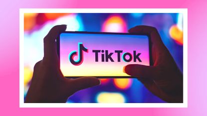 A silhouetted woman's hands holding a smartphone with the TikTok logo displayed on the screen/ in a pink and purple template