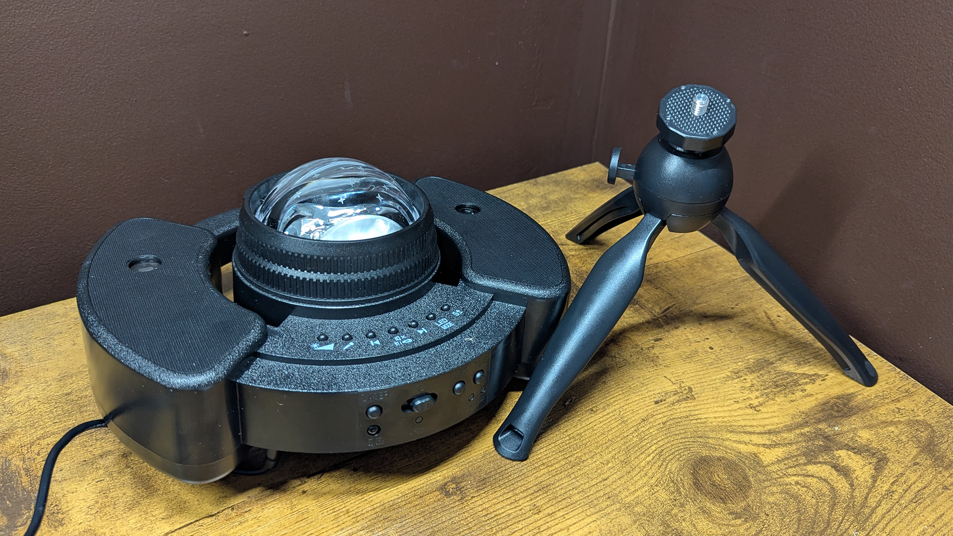 YgiTK Northern Galaxy Lights Projector next to its detached tripod during review. It is on a wooden table against a dark coloured wall