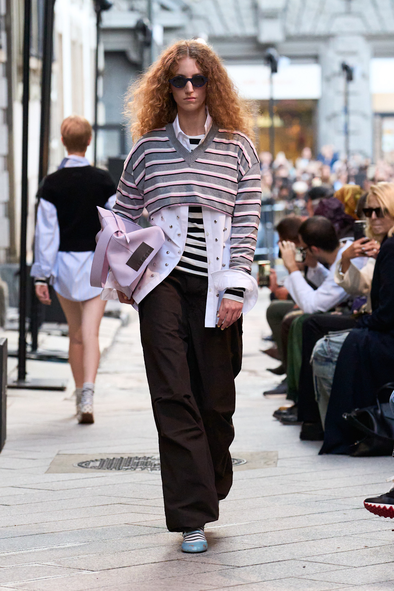 Layering staples for spring 2026 is shown in a photo from MSGM's runway show, with a model walking while wearing a gray-striped V-neck sweater layered over an unbuttoned oversized Oxford shirt, with a black-and-white striped t-shirt peeking out underneath. The look is also styled with brown trousers, an oversized pink bag, striped socks, black sunglasses, and blue Mary Jane flats 