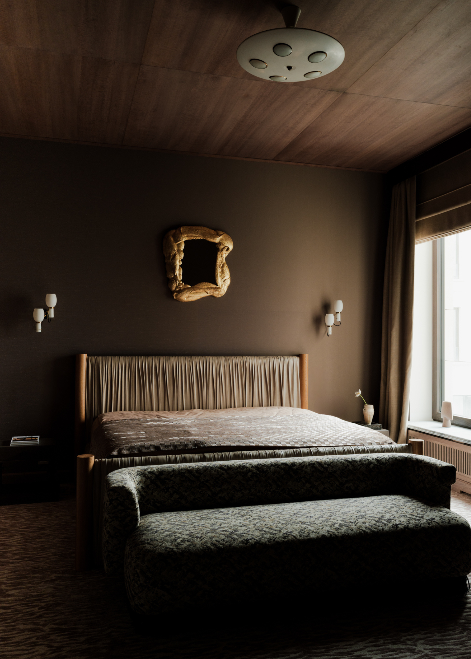 A dark chocolate brown bedroom with white wall sconces, a moulded wood framed mirror, a headboard with gathered fabric, a patterned bench, and a side table with a vase