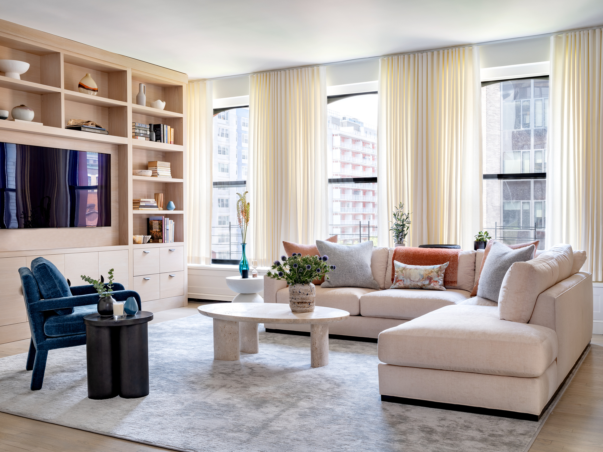 apartment living room layout with wall of windows, a built-in cabinet with TV, a stone coffee table, L-shaped sofa, and blue armchair with a side table