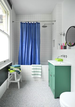 white bathroom with blue shower curtain, green vanity, white tiled walls, grey flecked vinyl, stool, towels