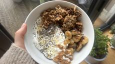 Hand holding bowl of yoghurt, granola, fried banana and peanut butter seen from above
