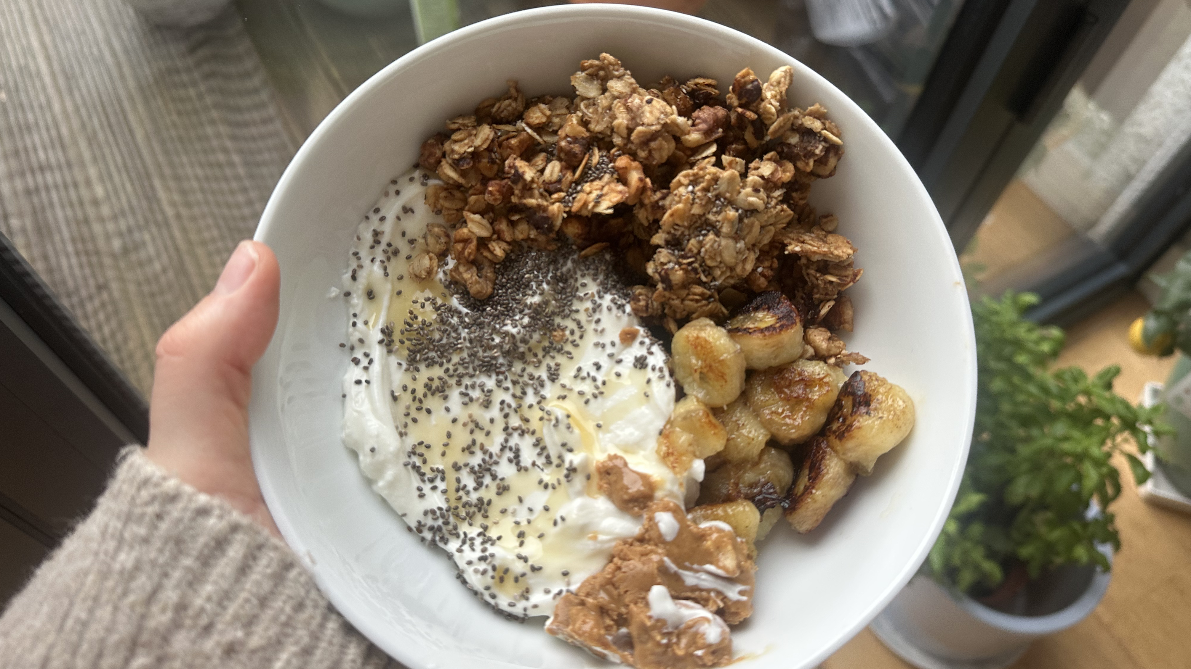 Hand holding bowl of yoghurt, granola, fried banana and peanut butter seen from above