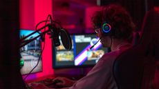 Young man playing video games on a desktop computer in a dimly lit room, with glowing headphones.