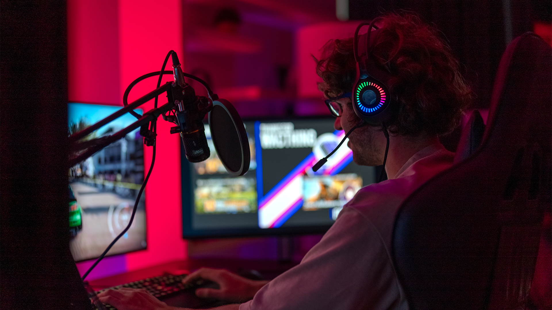 Young man playing video games on a desktop computer in a dimly lit room, with glowing headphones.