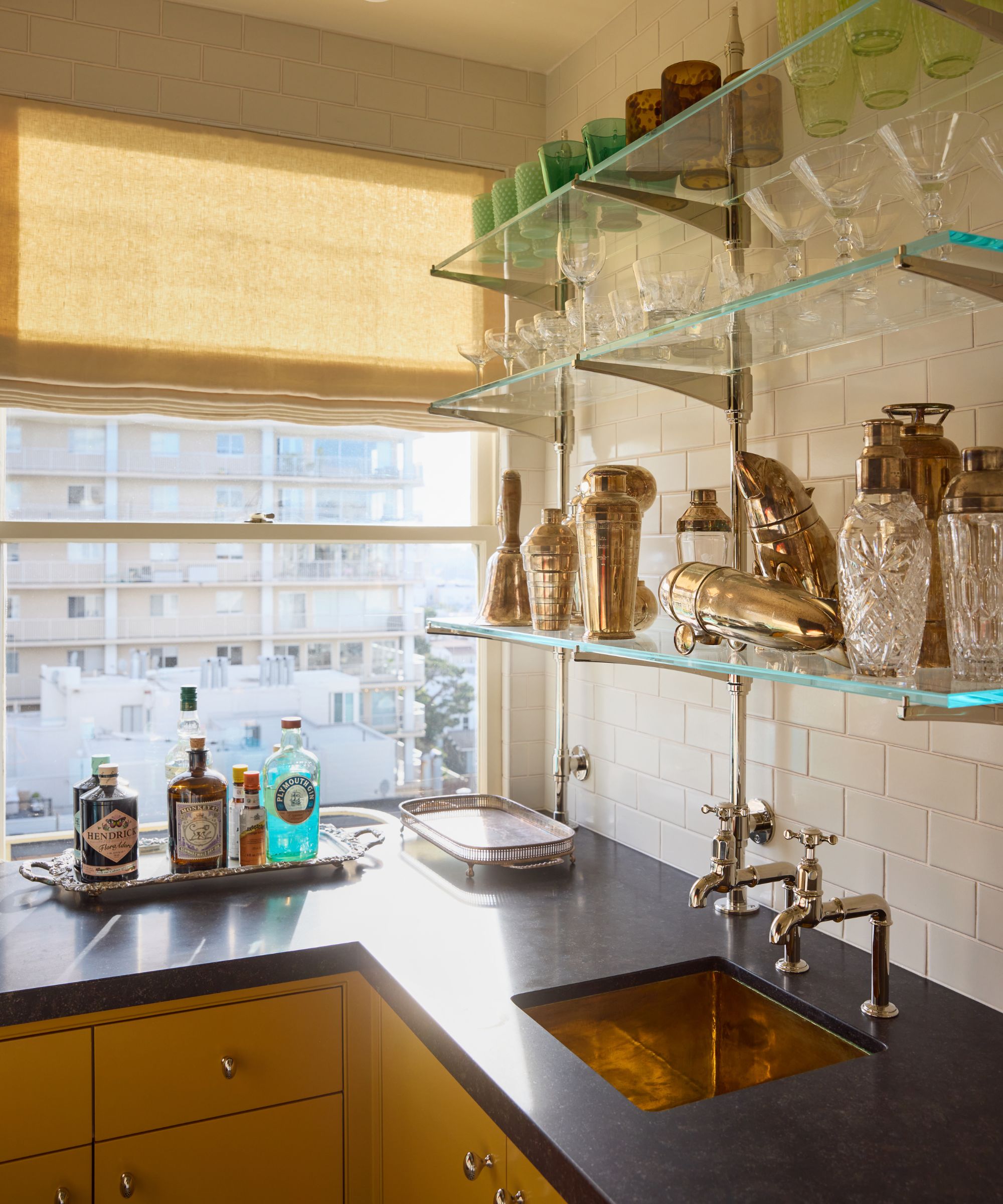 a small deep yellow colored home bar with dark stone counters, brass sink, chrome and glass open shelving styled with vintage cocktail equipment