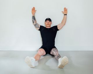 Coach Corey Perkins performs a wall slide. He is sitting on the floor, with his back resting on a wal and his feet straight out in front of him. His arms are held aloft in a 'u' shape.