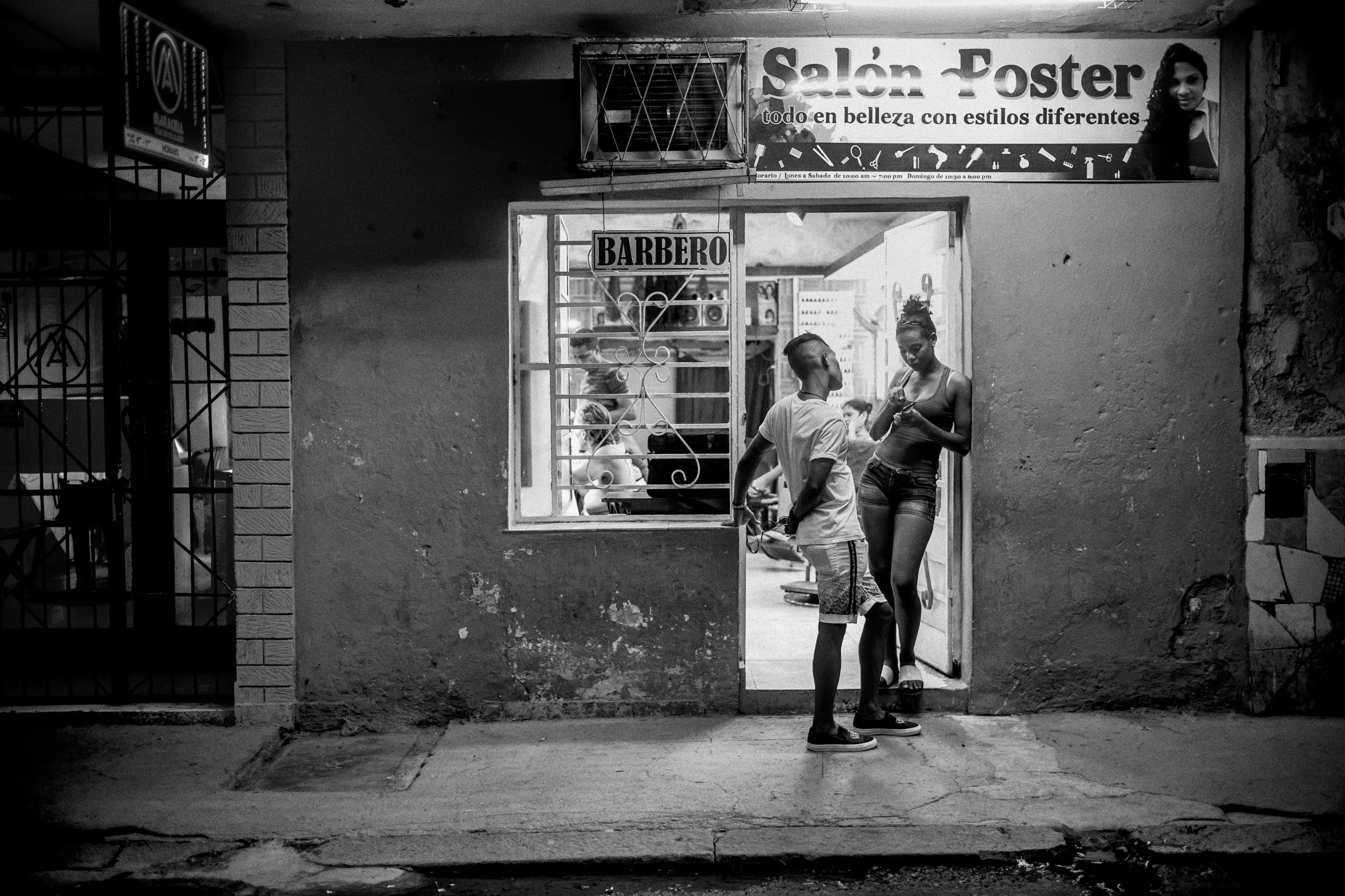 Black and white night shot of a young couple standing outside a hair salon in Havana, its illuminated sign reading "Sal&amp;oacute;n Foster, todo en belleza con estilos diferentes."