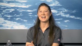 a smiling woman in a gray shirt sits at a table with a large photo of earth from space in the background